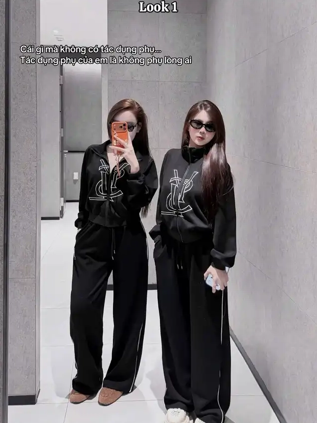 Hai cô gái mặc bộ đồ thể thao màu đen đồng điệu với logo trắng đeo kính râm chụp ảnh selfie trong gương