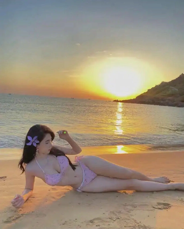 Một cô gái mặc bikini màu hồng nằm nghiêng trên bãi cát nhìn về phía mặt trời lặn trên biển với bông hoa cài trên tóc